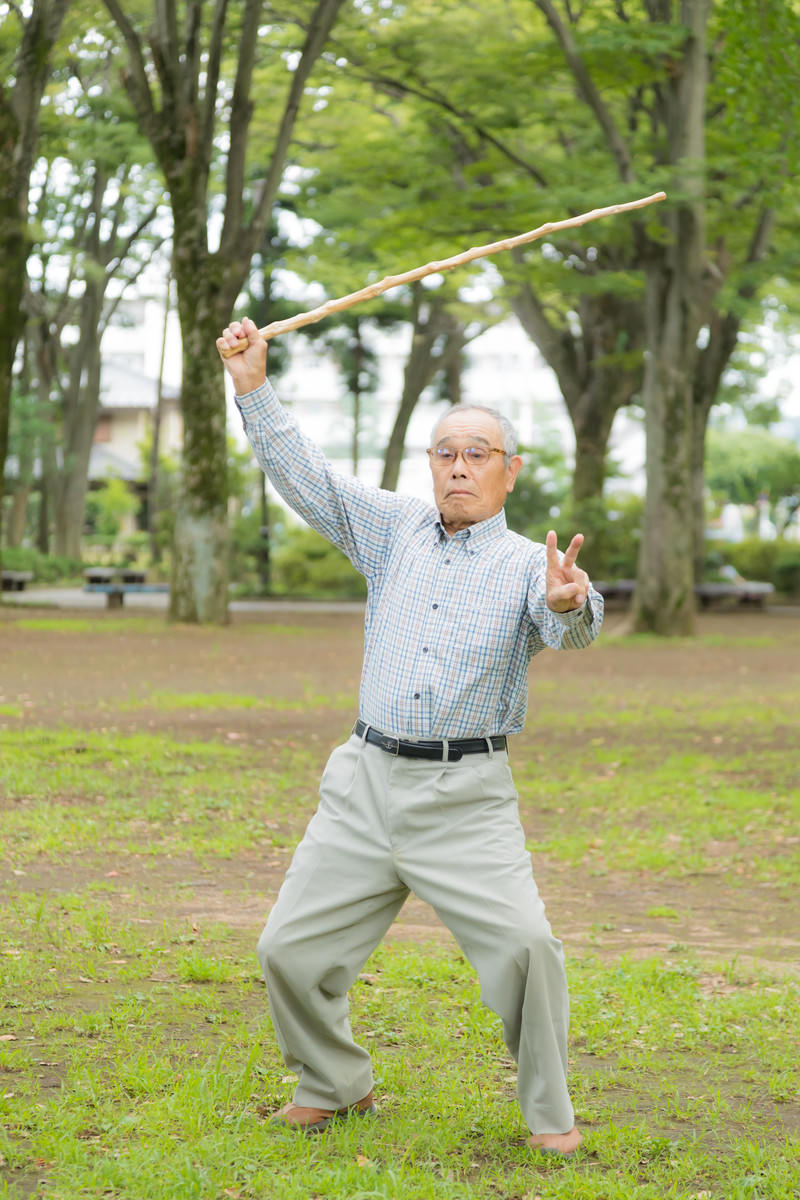 公園で棒を振り上げて笑顔でポーズを取る白髪のお爺さん［モデル：まーちゃん］