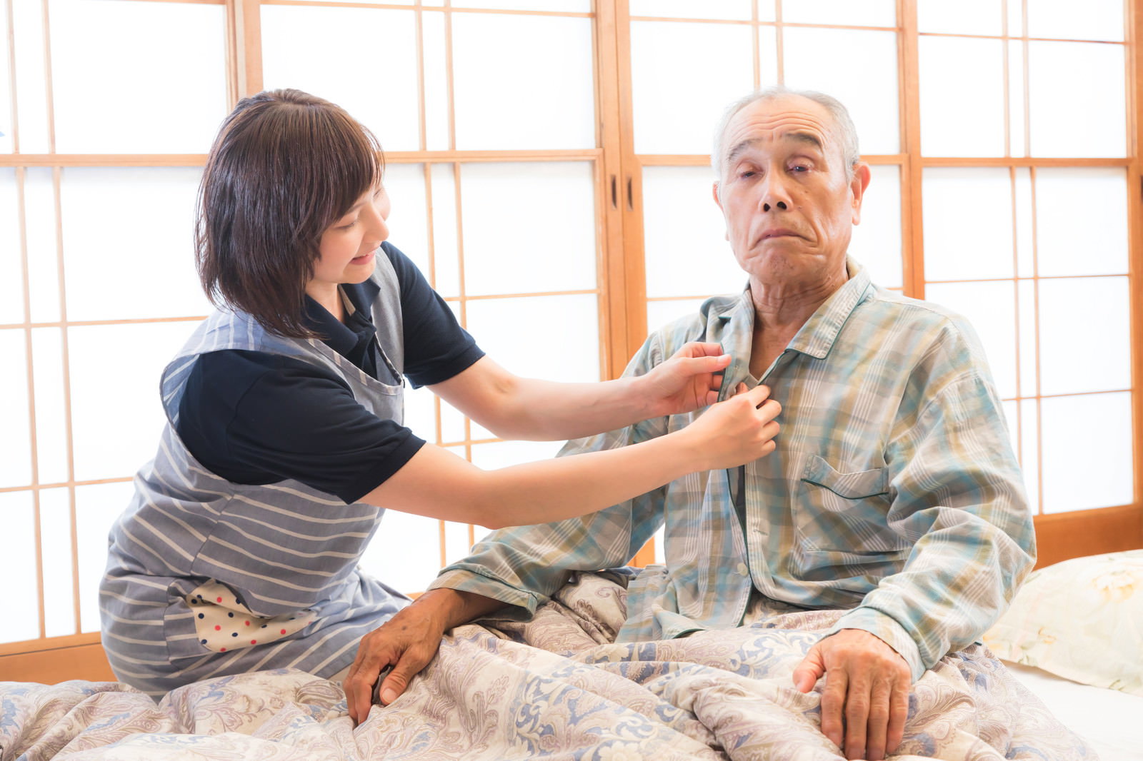 ベッドで横になった高齢男性のパジャマのボタンを留める女性介護士［モデル：まーちゃん yumiko］