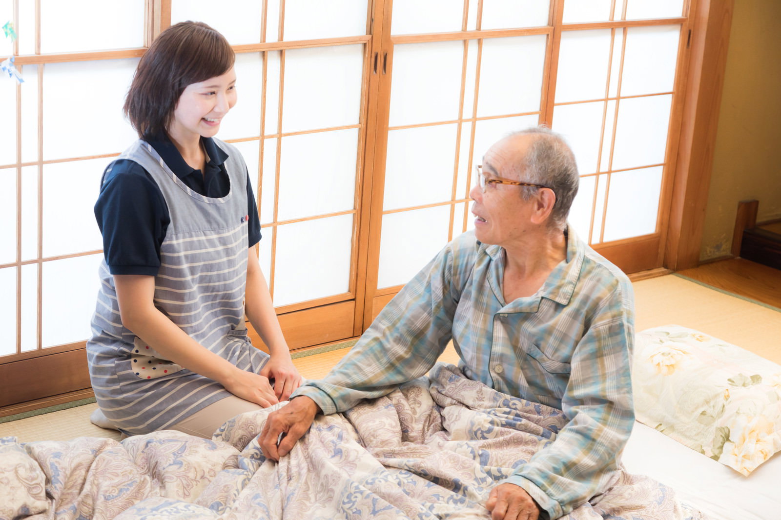 布団に横になったお爺さんと女性介護士が穏やかに会話している出張介護の風景［モデル：まーちゃん yumiko］