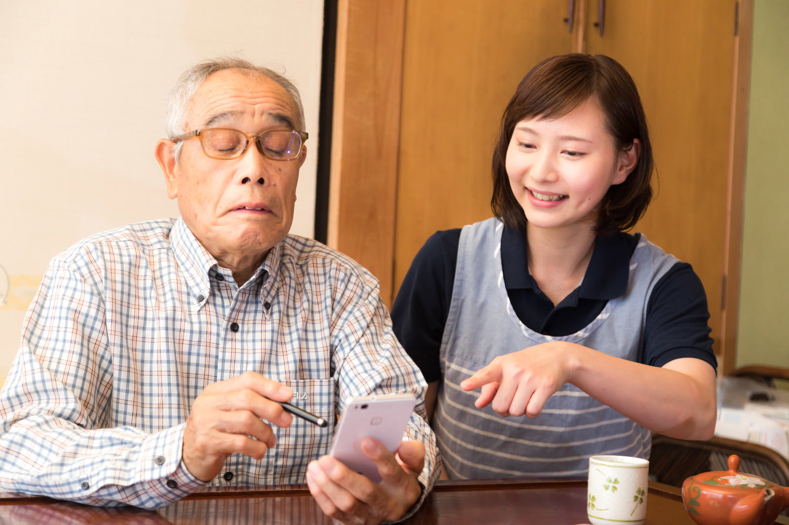 女性介護士からスマホの使い方を教わる眼鏡をかけた高齢男性［モデル：まーちゃん yumiko］