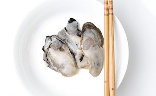 箸で取る皿に盛り付けた牡蠣の身、竹製の取り箸が添えられた白い皿
