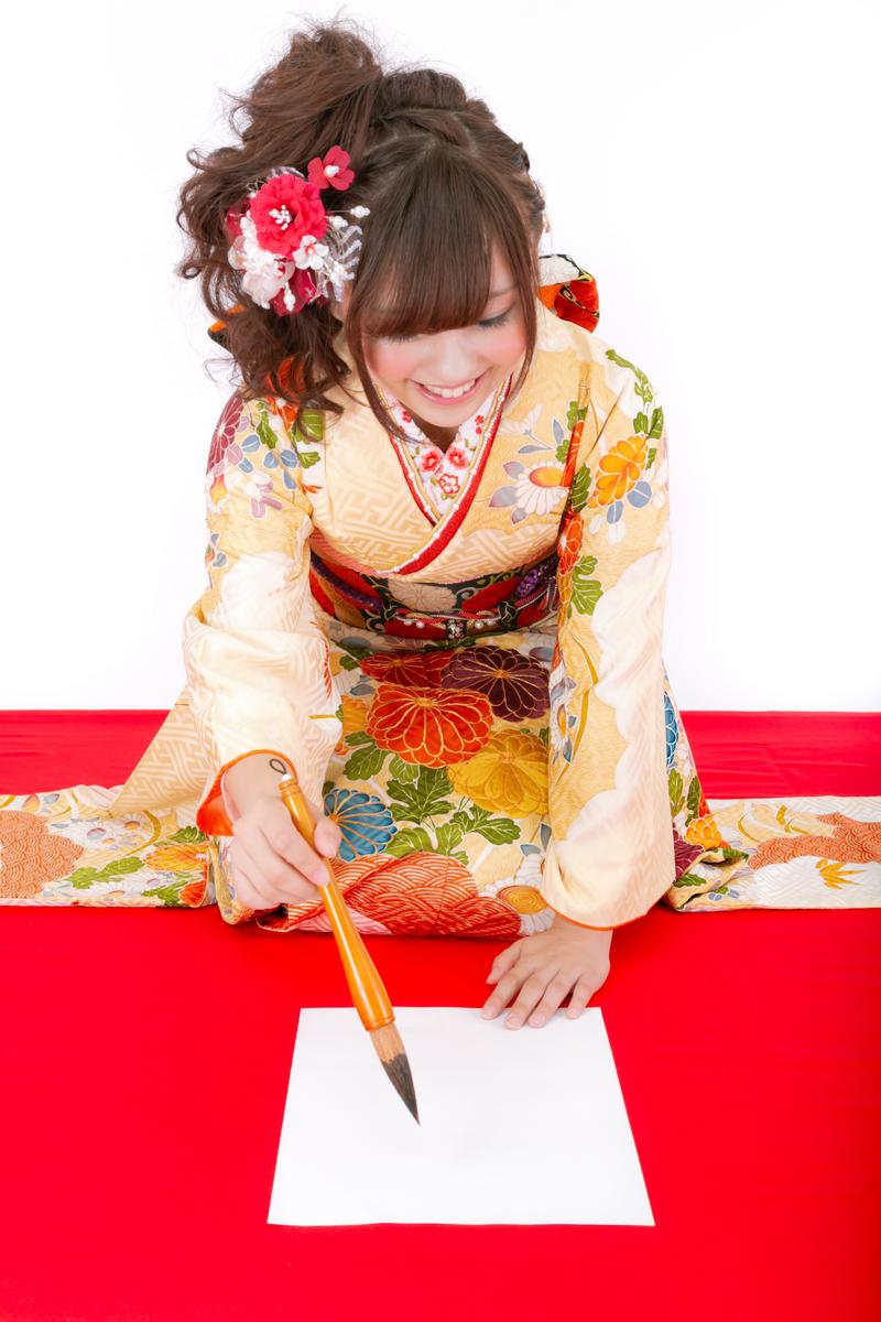 振袖を着た女性が赤いテーブルの白い紙に書き初めをしている［モデル：河村友歌］