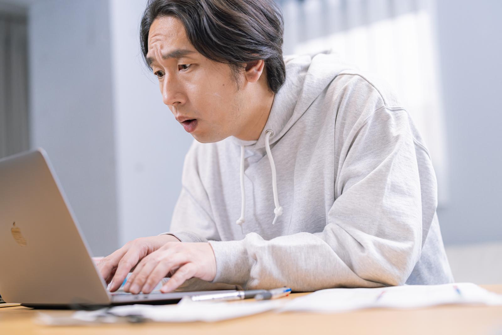 グレーのパーカーの男性がパソコン画面を前に口を開けた驚きの表情で、キーボードに手を置いている［モデル：大川竜弥］