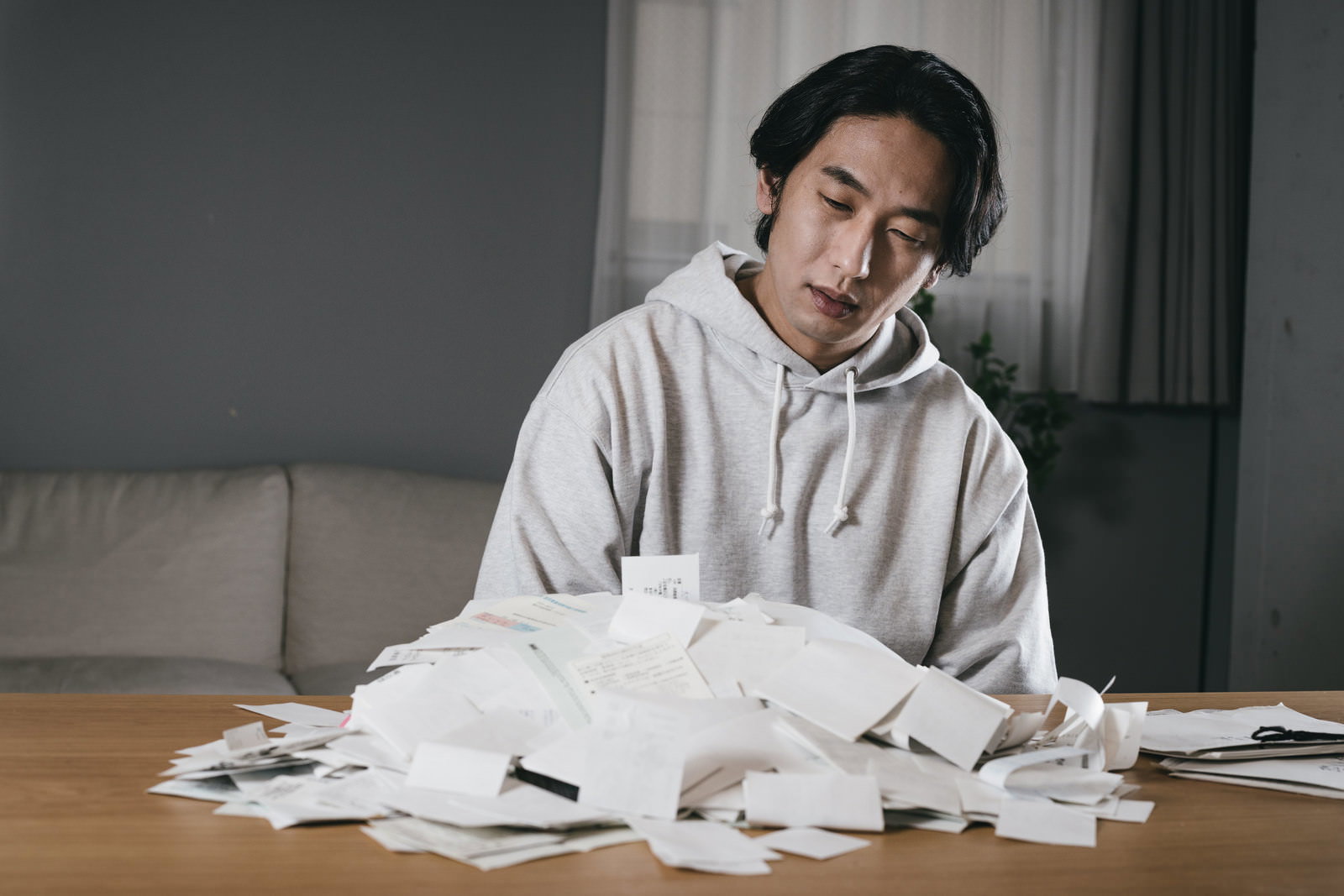 グレーのパーカーを着た男性がテーブルに積み重ねられた領収書を見つめている［モデル：大川竜弥］