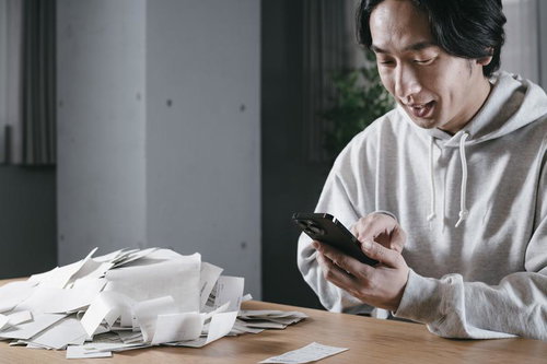 テーブルに散乱した大量の領収書を前にスマートフォンで作業する男性