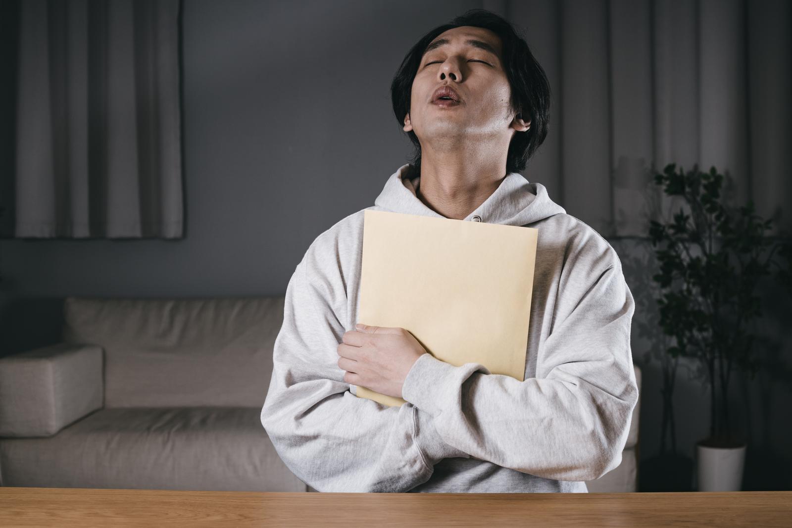 グレーのパーカーを着た男性が黄白色の封筒を胸に抱きしめて顔を上げている［モデル：大川竜弥］