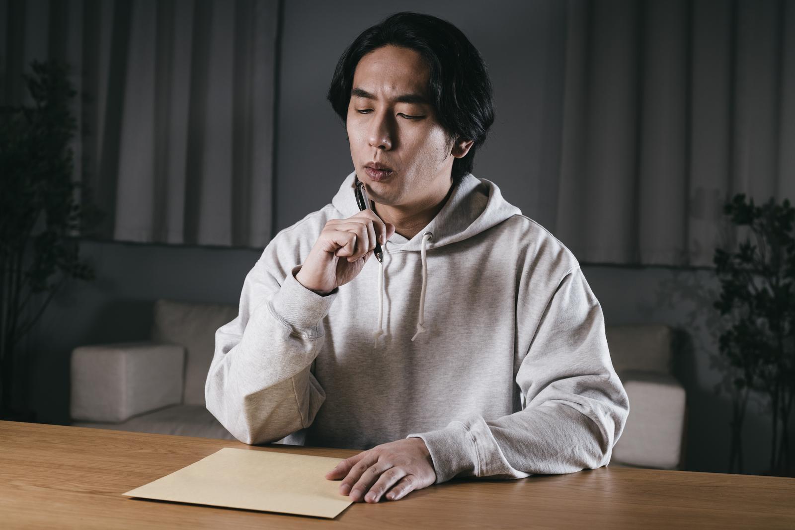 ペンを持って白い用紙を前に悩む、ベージュのパーカー姿の男性［モデル：大川竜弥］