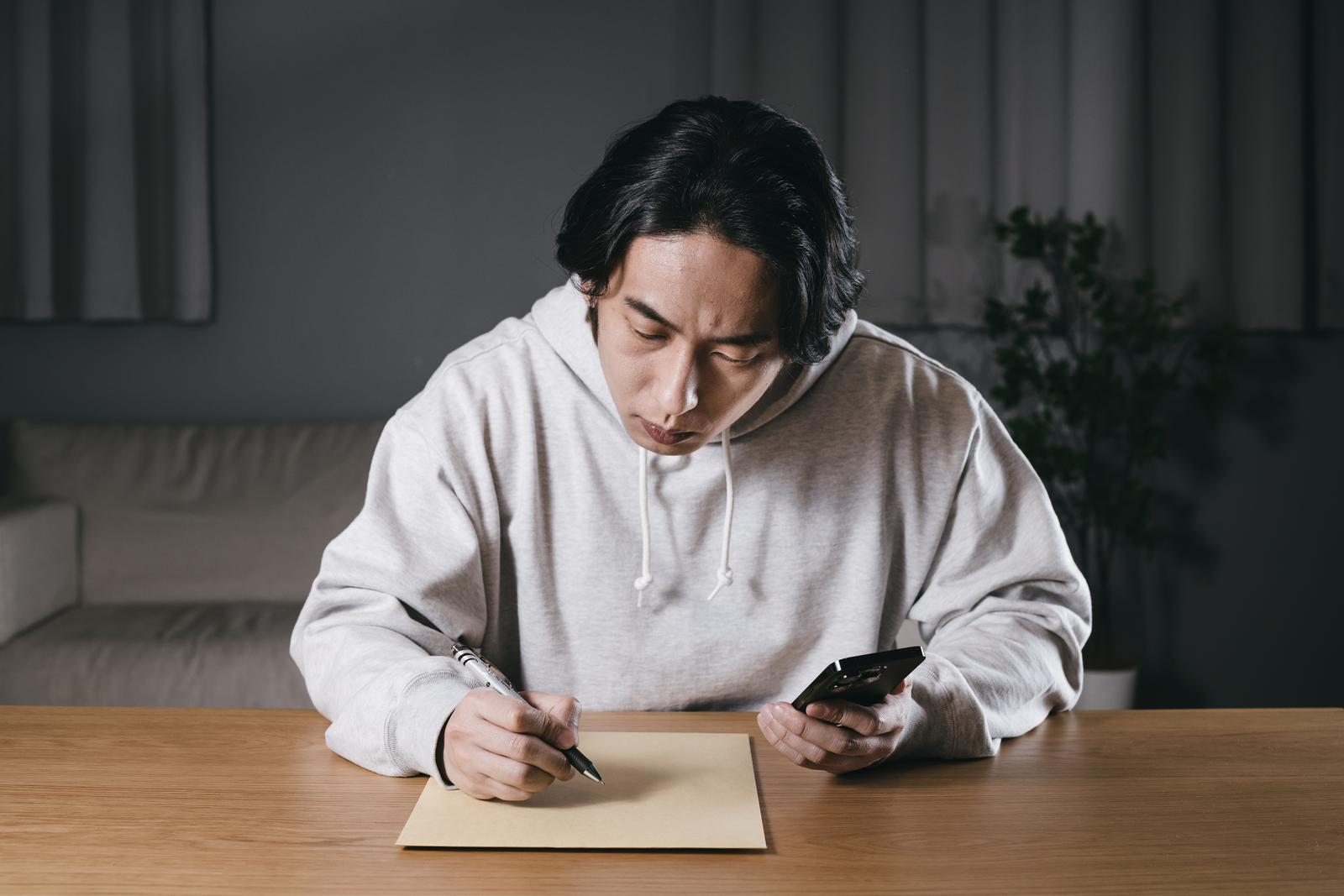 スマートフォンを見ながら宛先を手書きする男性［モデル：大川竜弥］
