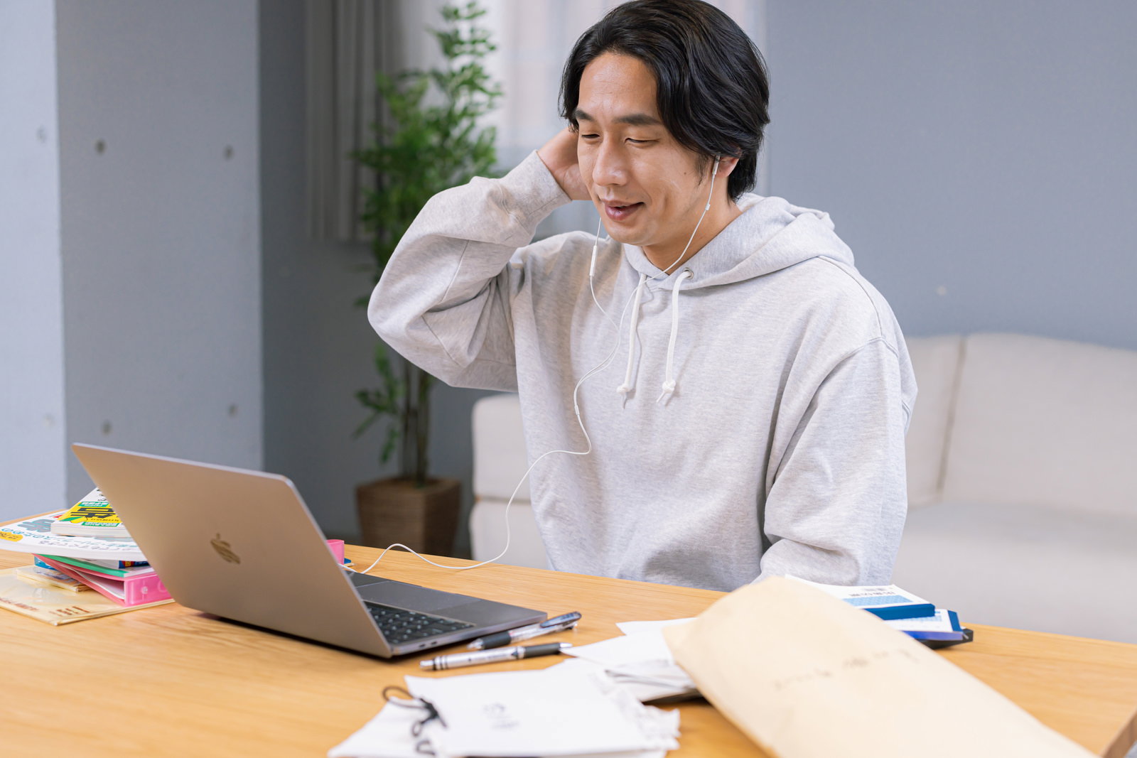 グレーのパーカーを着た男性がノートパソコンでオンライン会議をしながら書類を確認している［モデル：大川竜弥］