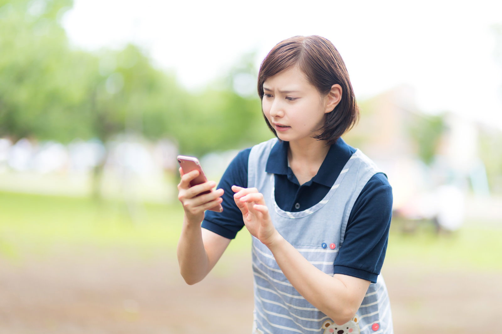 スマホを見ながら困った表情を浮かべるエプロン姿の女性介護士［モデル：yumiko］