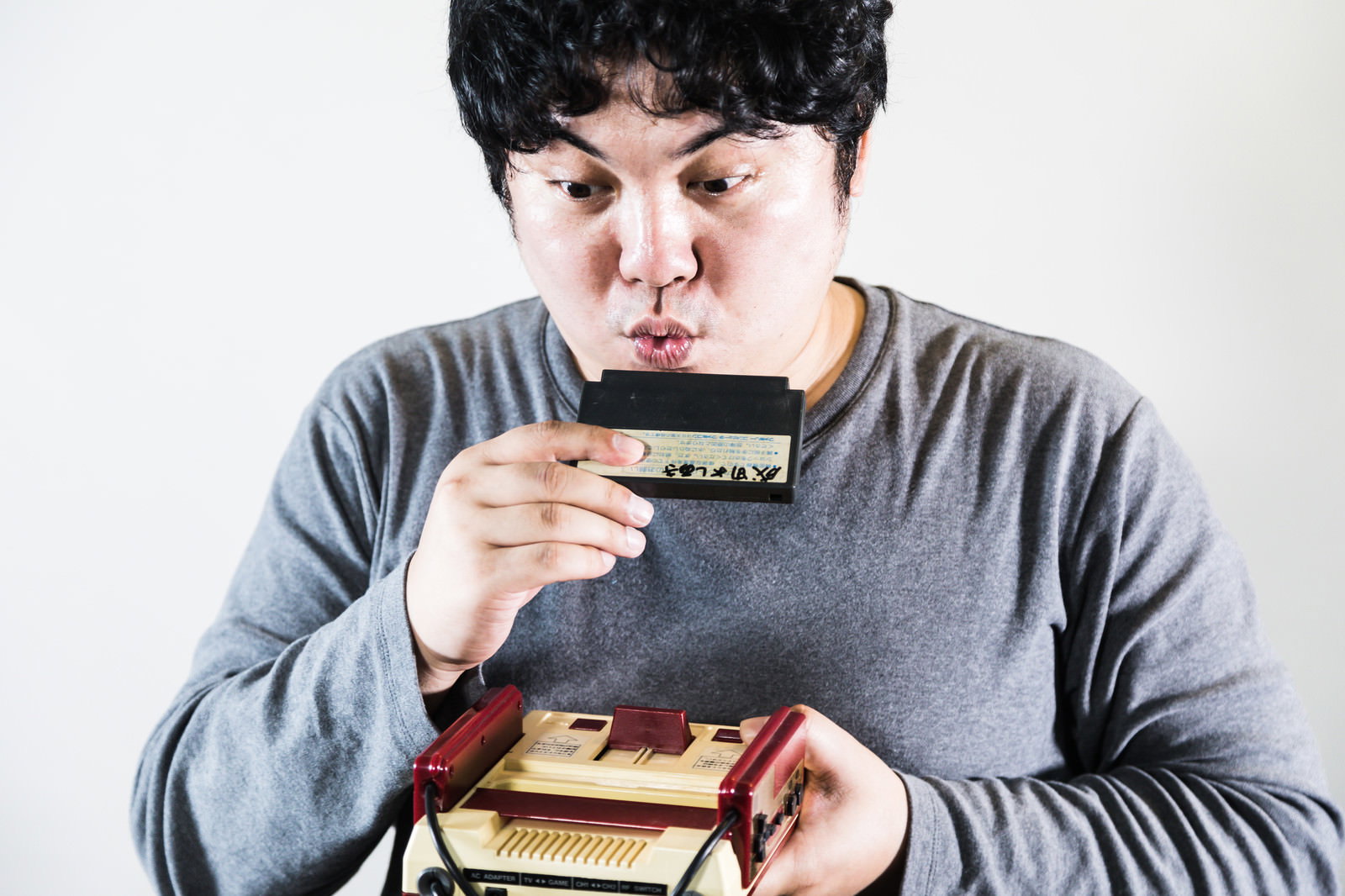 ファミコンカセットに息を吹きかけるグレー服の男性［モデル：段田隼人］