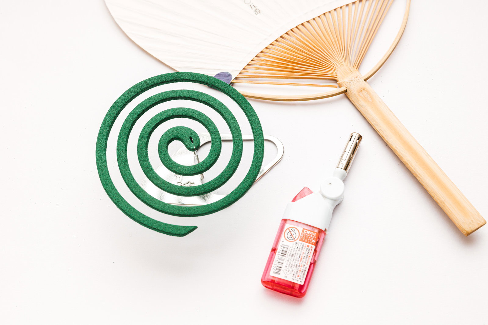 白い背景に置かれた緑色の蚊取り線香と竹製のうちわ、赤いライター