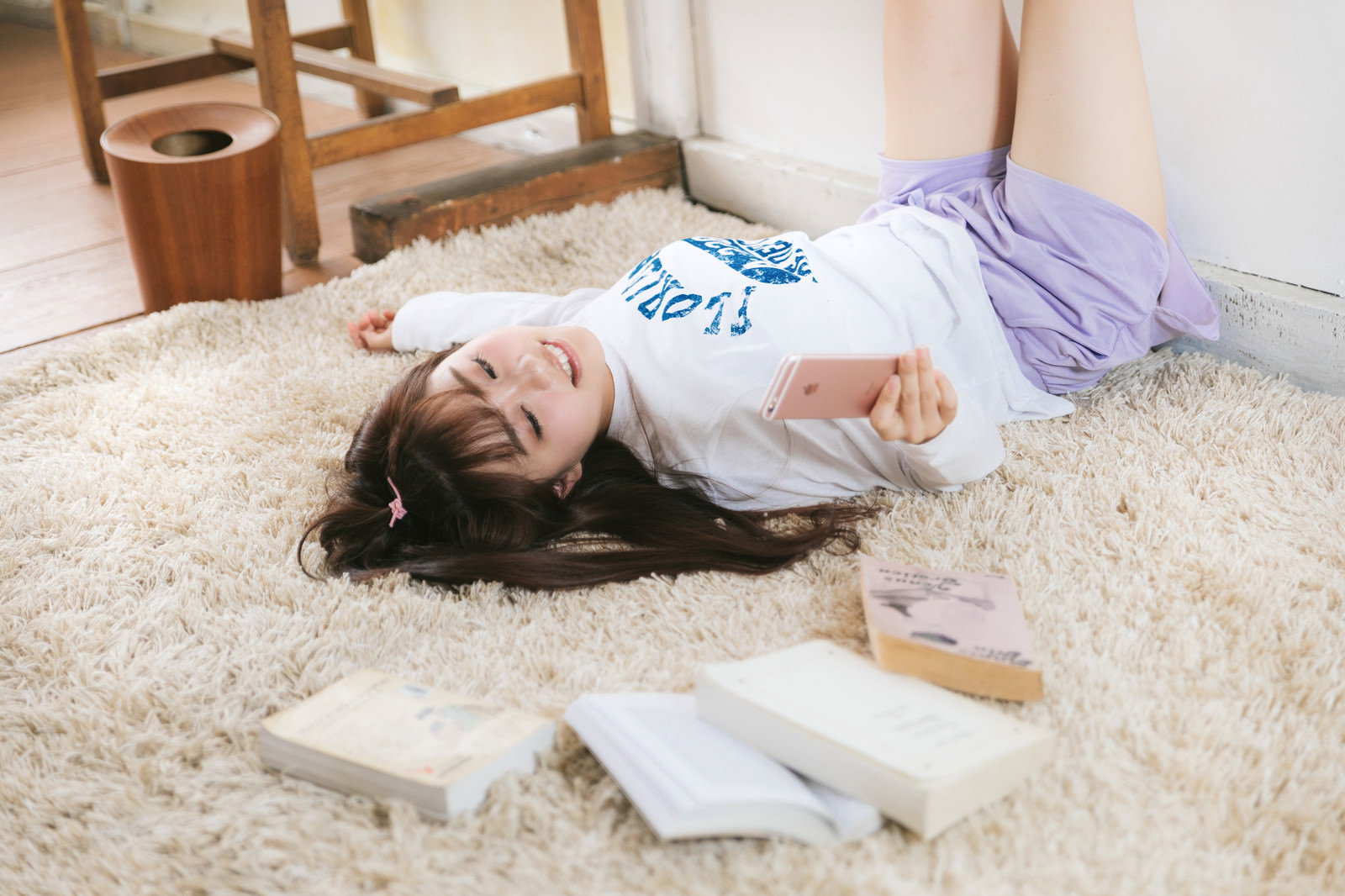 カーペットの上で部屋着を着た女性が本を置いてスマホを見ている［モデル：河村友歌］