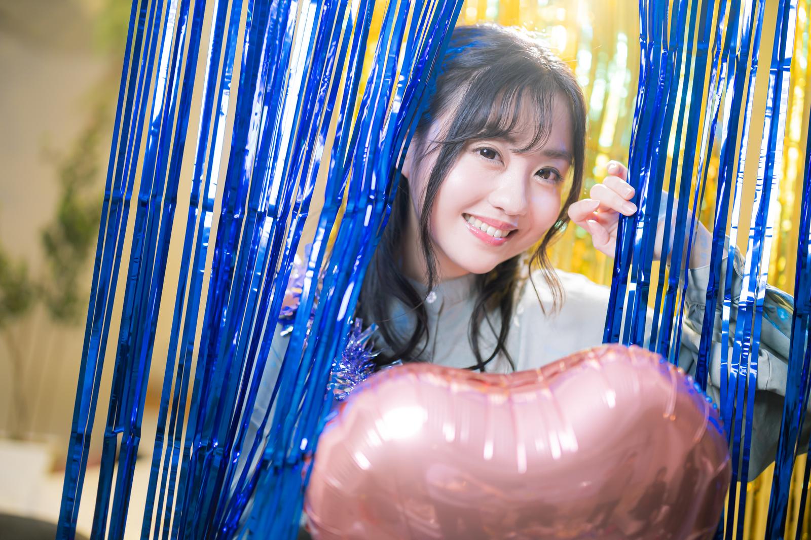 青と黄色の装飾に囲まれた女性がピンク色の風船を持って笑顔で顔を出している［モデル：河村友歌］