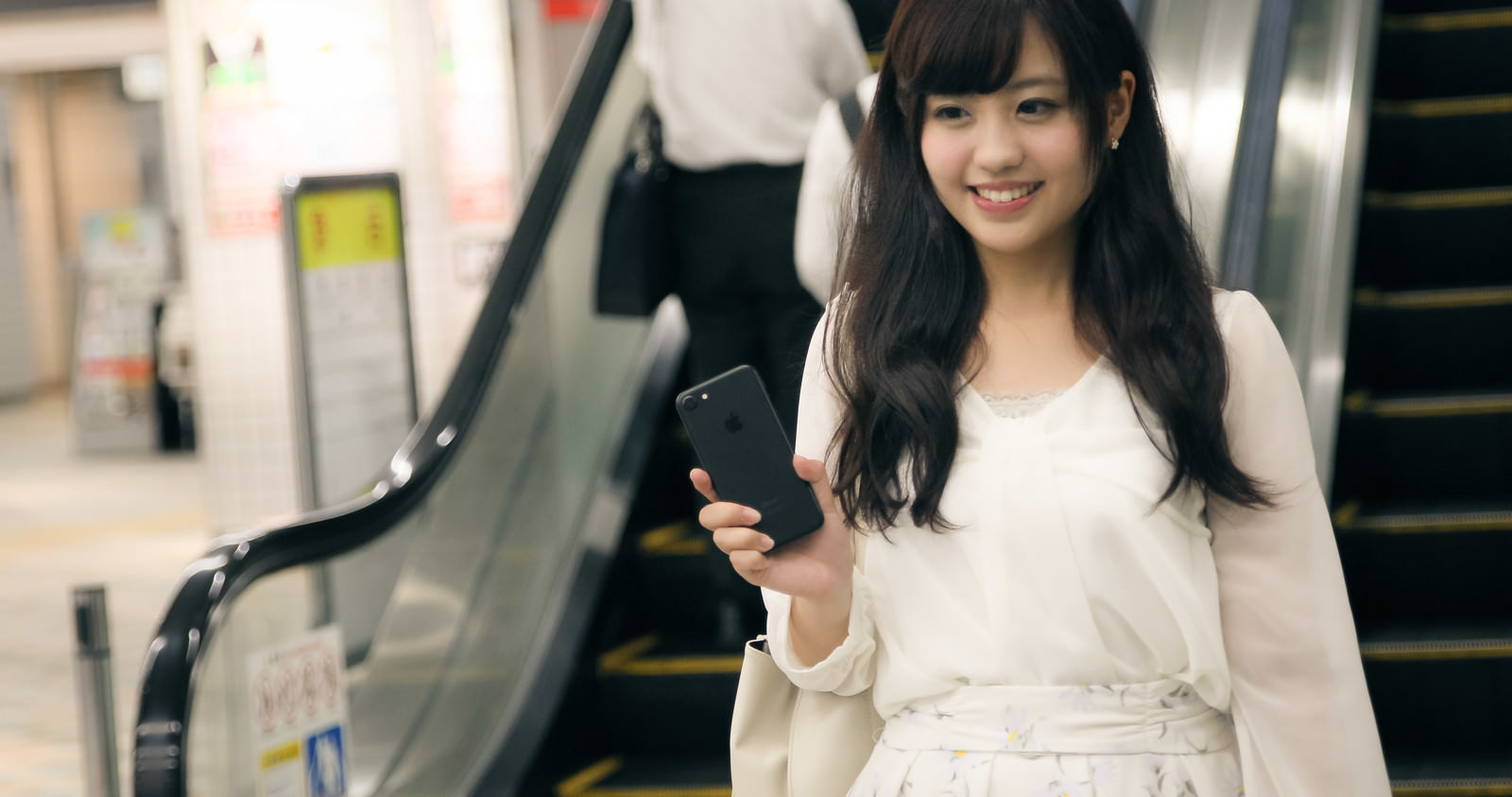 駅構内でiPhoneを手に持ち微笑む女性［モデル：河村友歌］