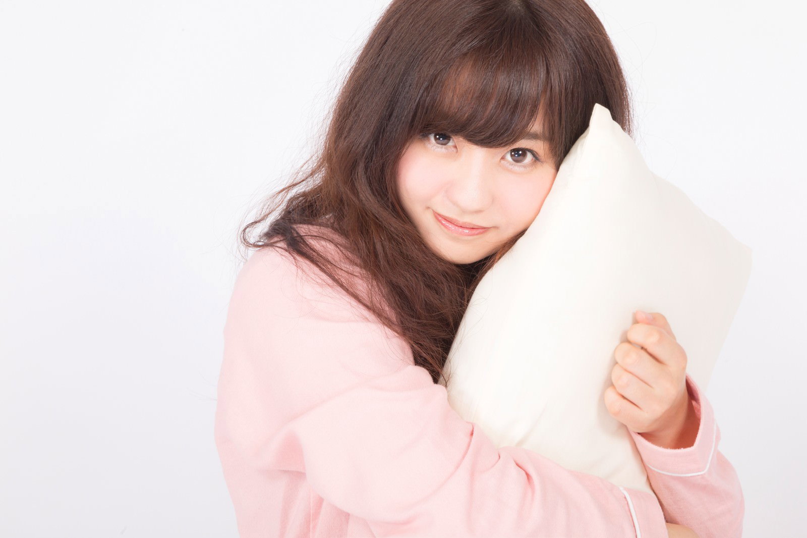 ピンクのパジャマを着た女性が白いまくらを抱きしめて上目遣いで見つめている［モデル：河村友歌］