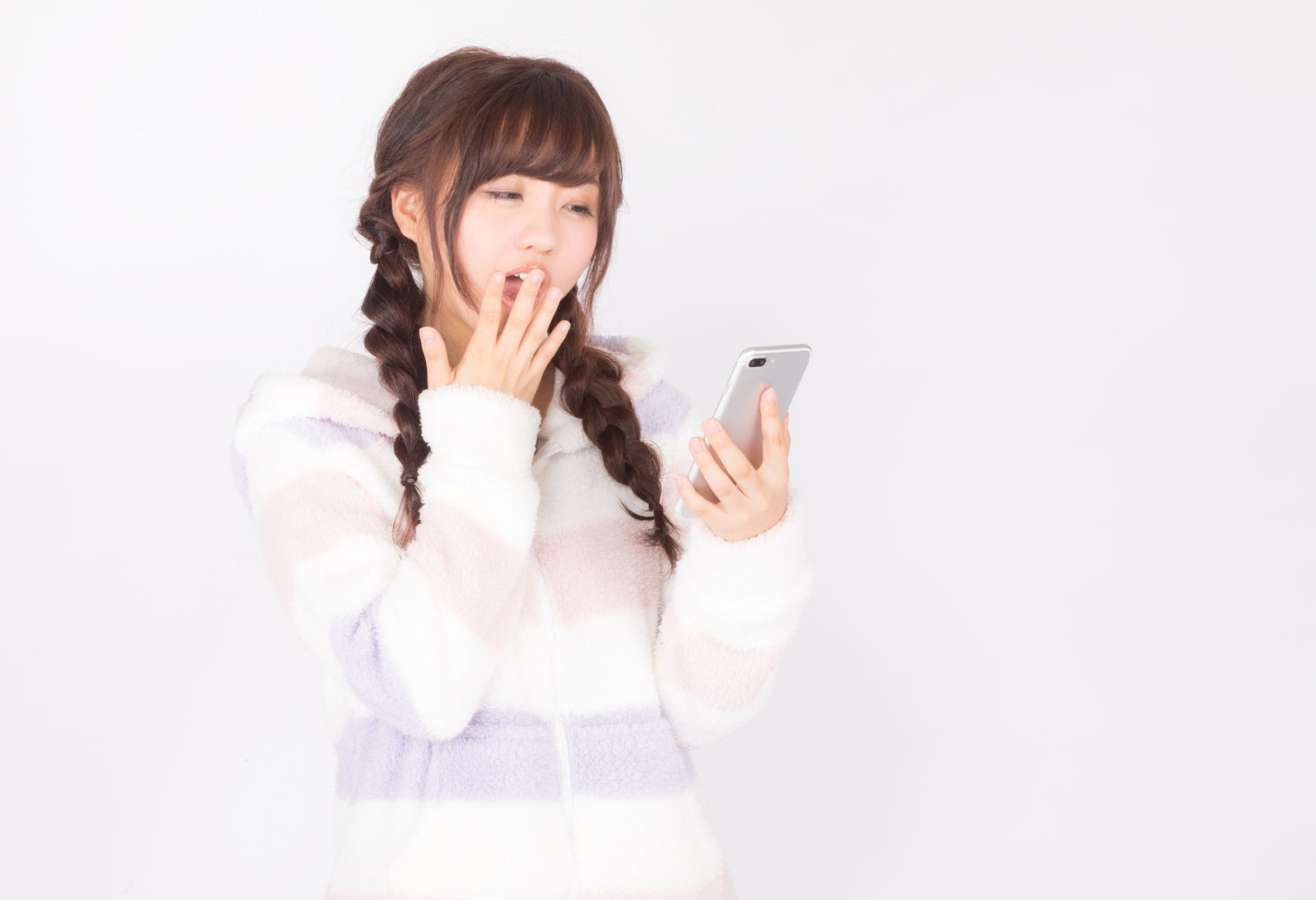 白いニット素材の服を着て、スマートフォンを持ちながらあくびをする寝不足の女性［モデル：河村友歌］