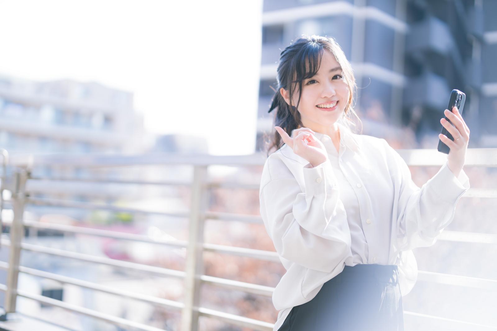 スマートフォンで自撮りする白シャツの笑顔の女性［モデル：河村友歌］