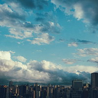 上空に晴れ間と雨雲が広がる都心の街並みと高層建築の写真