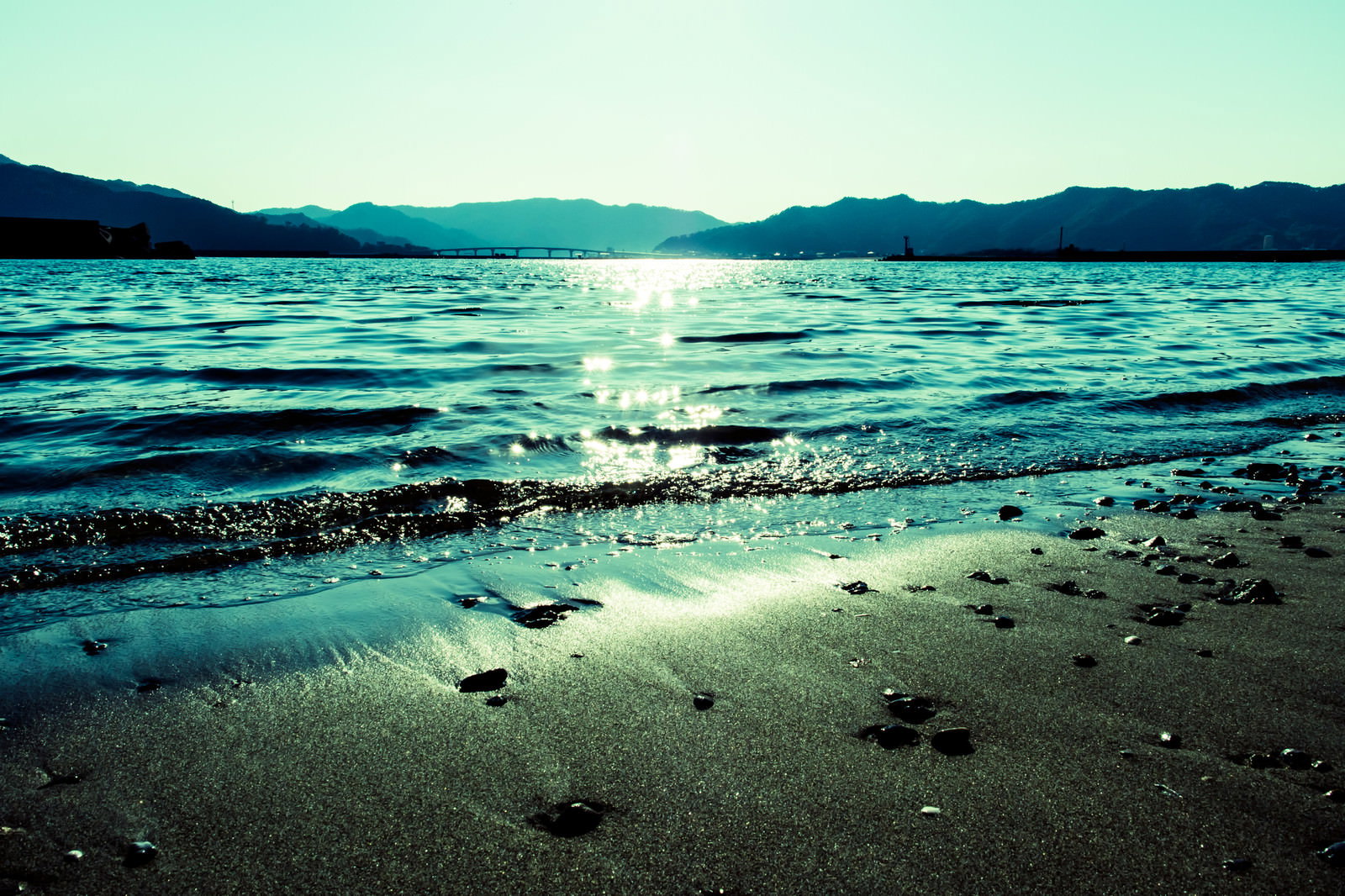光が反射する海と遠くの山々が見える砂浜の風景