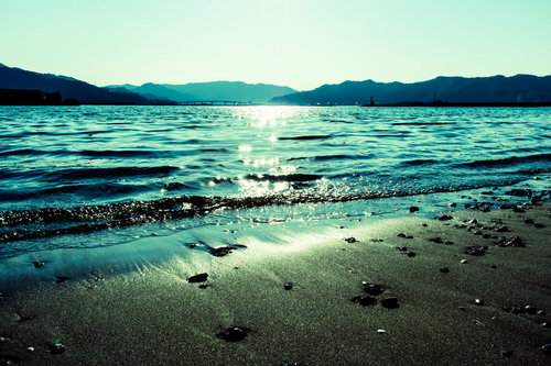 日の光が差し込む浜辺と遠くの山並み、波打ち際の風景