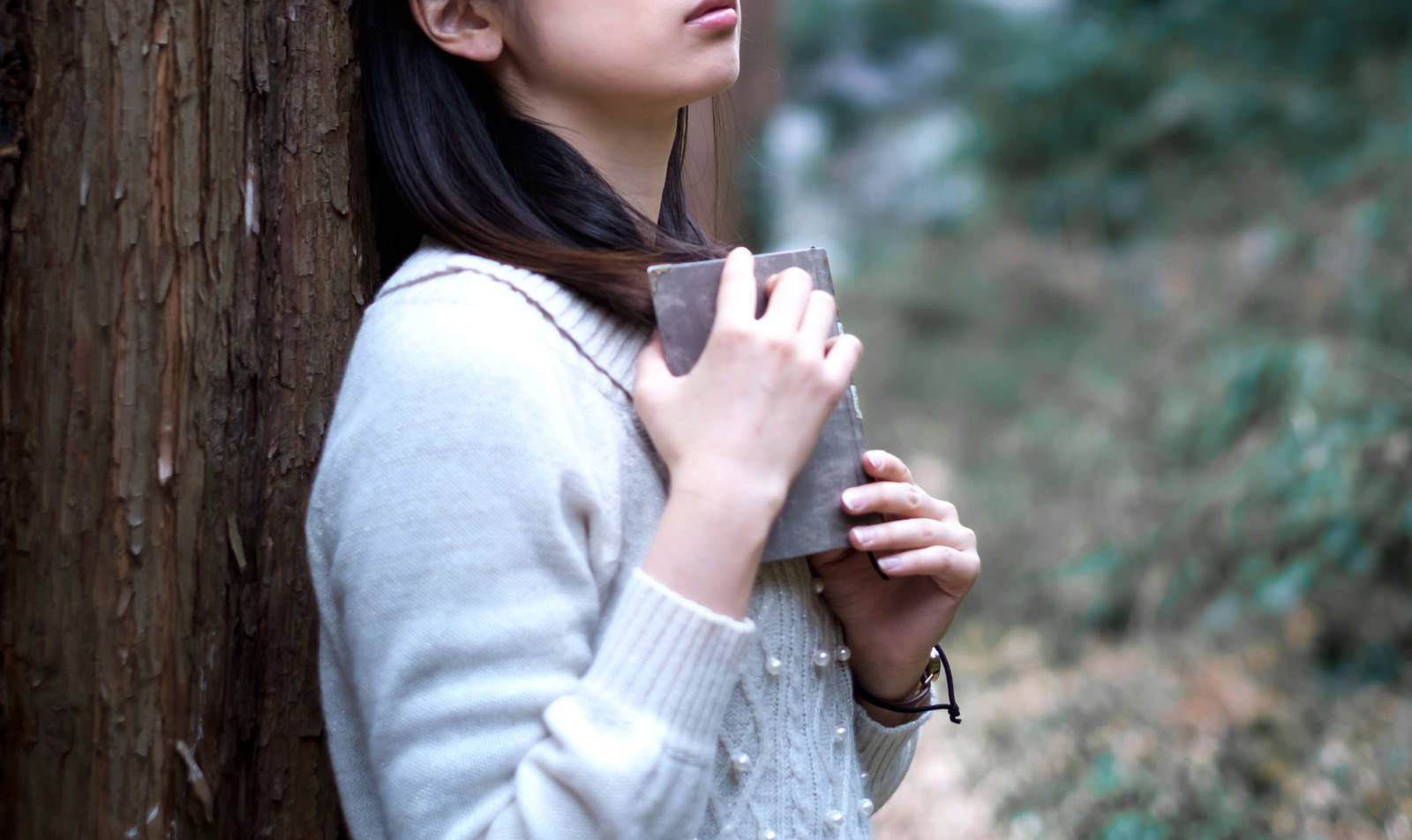 木の幹にもたれながら洋書を読んでいる白いトップスの女性