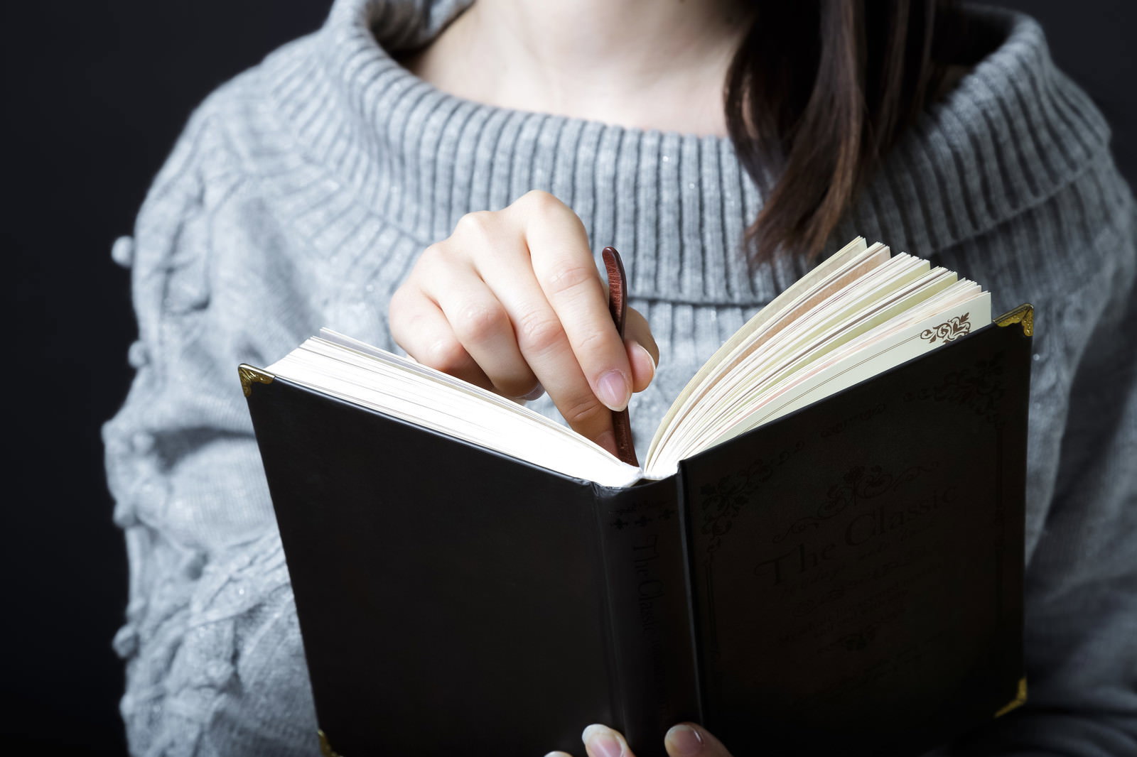 グレーのニットを着た女性が黒い洋書に栞を挟んでいる手元
