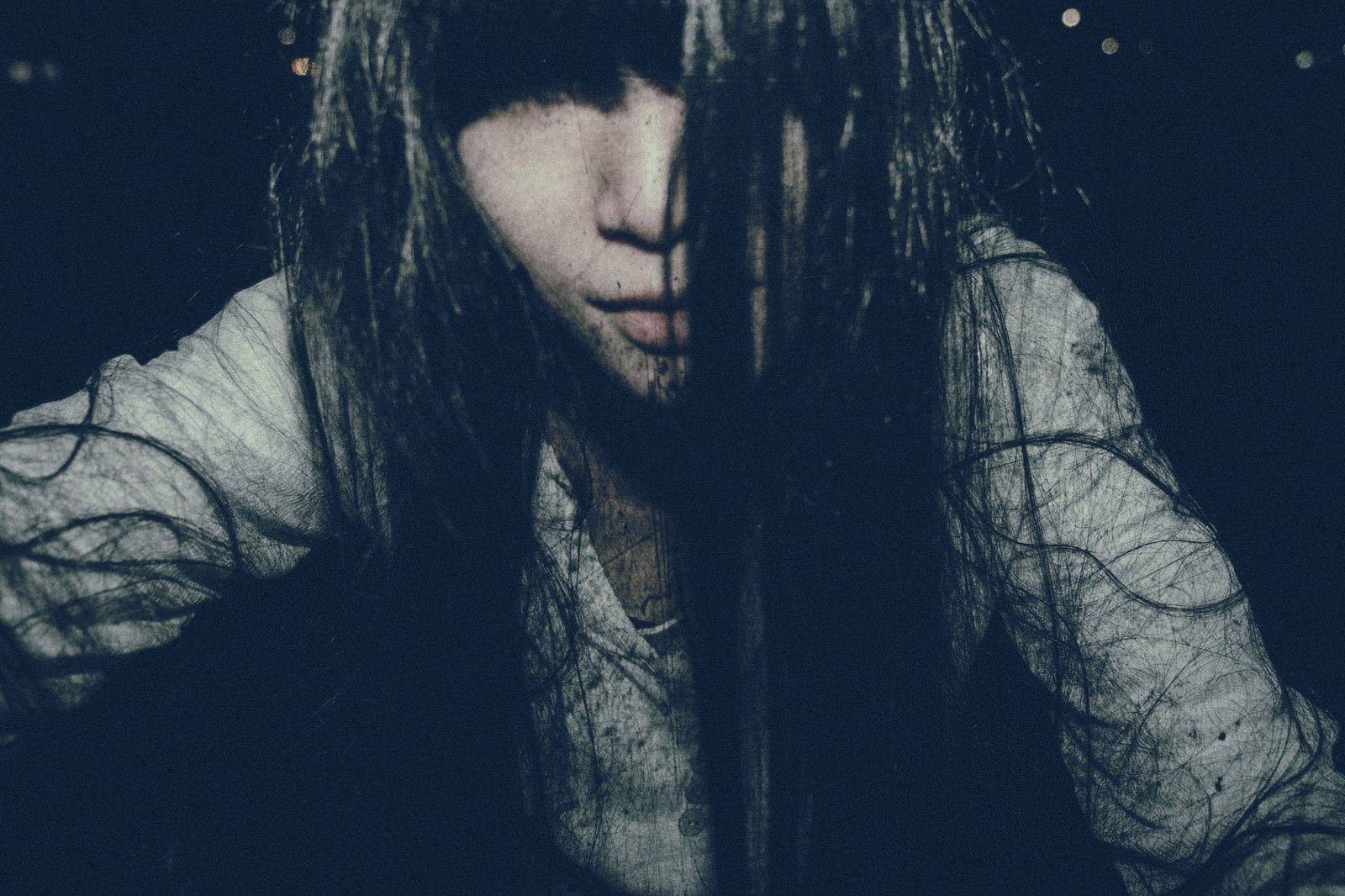 痛んだ長い黒髪で顔を覆った女性が暗闇の中にいる様子［モデル：緋真煉］