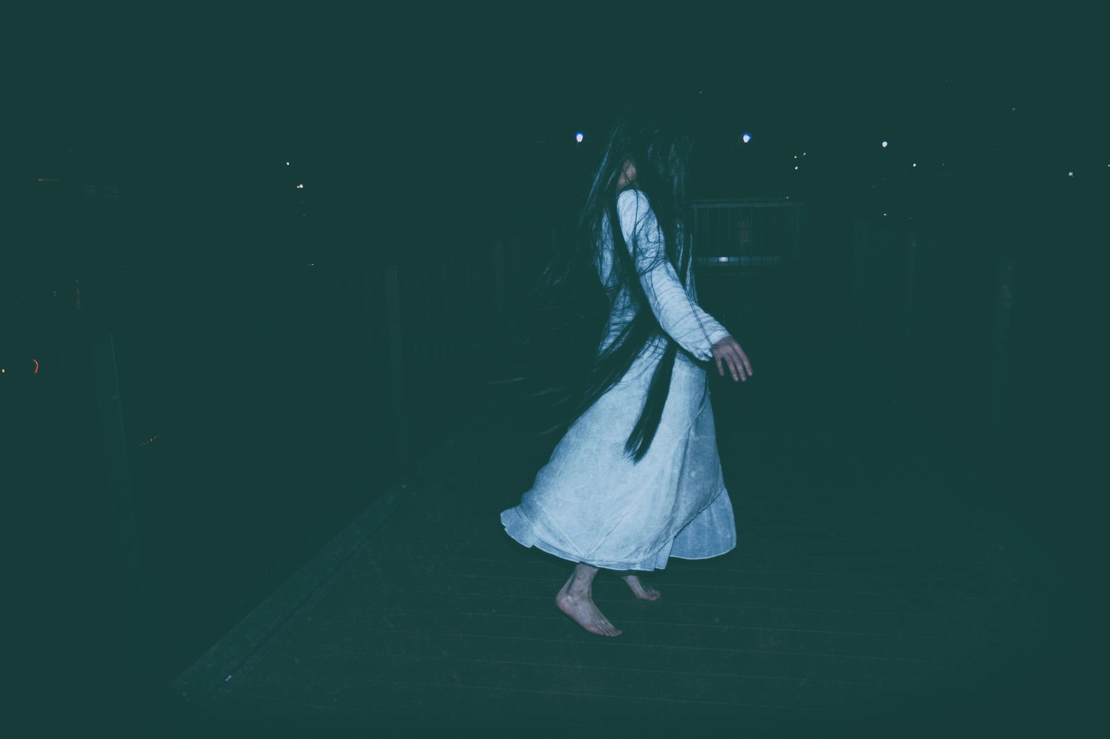 暗い背景の中に立つ白いワンピースを着た髪の長い女性［モデル：緋真煉］