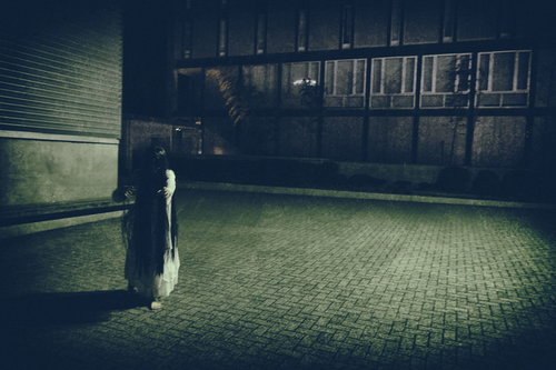 薄暗い石畳の広場へゆっくり近づいてくる白い服の女性の亡霊