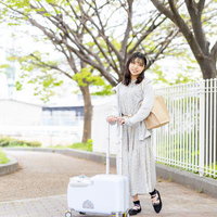 白いキャリーケースを持って新緑の歩道を歩く女性の写真