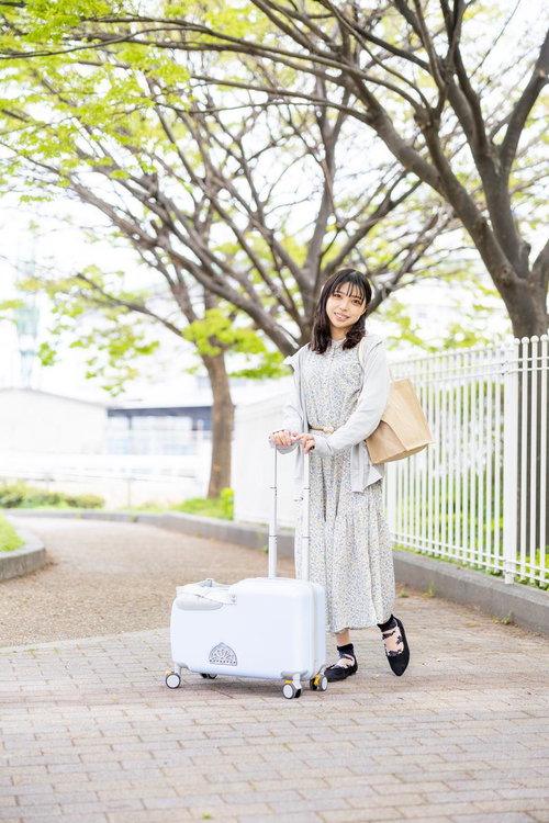 白いキャリーケースを持って新緑の歩道を歩く女性