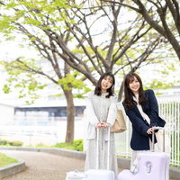 白いキャリーケースを持つ女性2人の新緑の並木道での女子旅シーンの写真