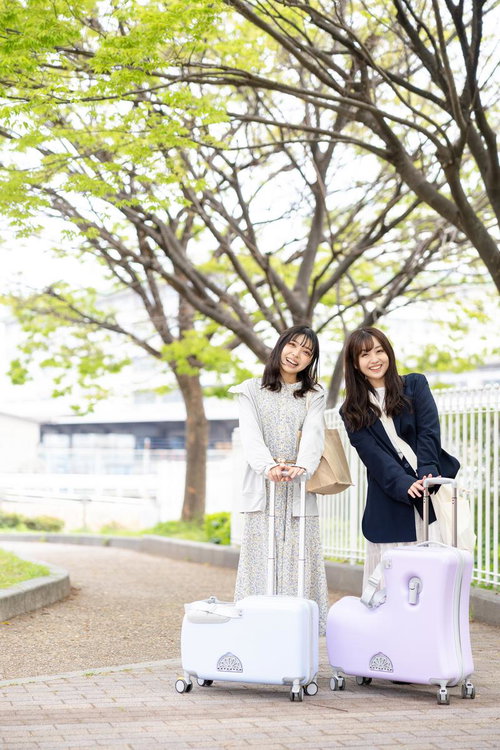 白いキャリーケースを持つ女性2人の新緑の並木道での女子旅シーン