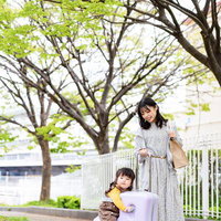 新緑の街路樹の下で紫のキャリーケースに座る子どもと母親の写真