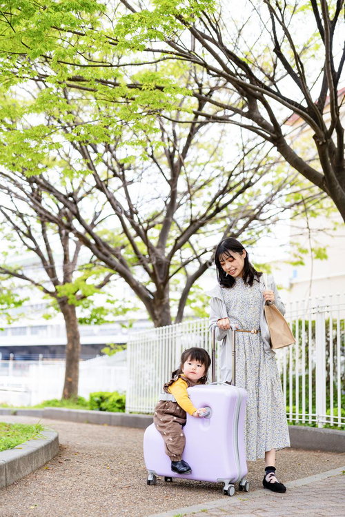 新緑の街路樹の下で紫のキャリーケースに座る子どもと母親