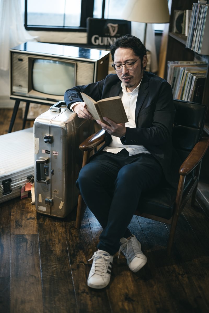 キャリーケースに寄りかかって洋書を読むスーツ姿の男性［モデル：ゆうせい］