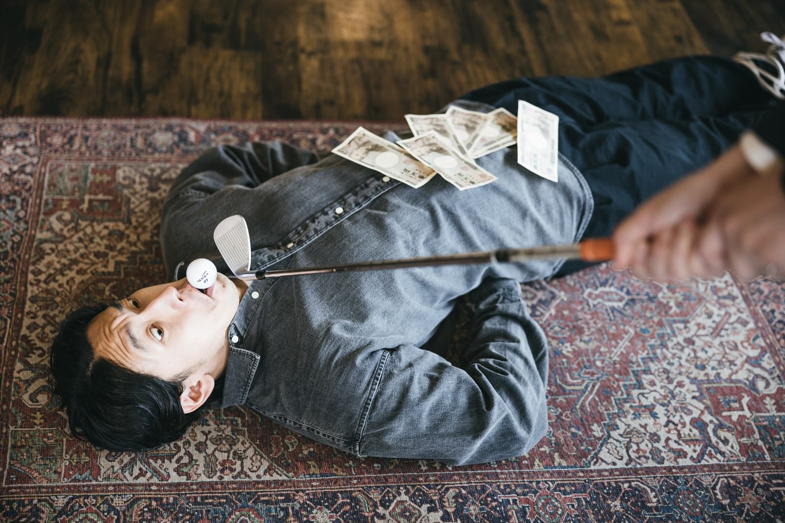 木製の床に倒れ込んだグレーのスウェット姿の男性と散らばった一万円札［モデル：大川竜弥］