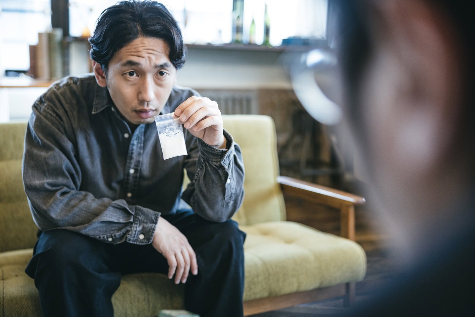 ソファに座りながら白い粉について尋問する刑事の男性［モデル：大川竜弥］