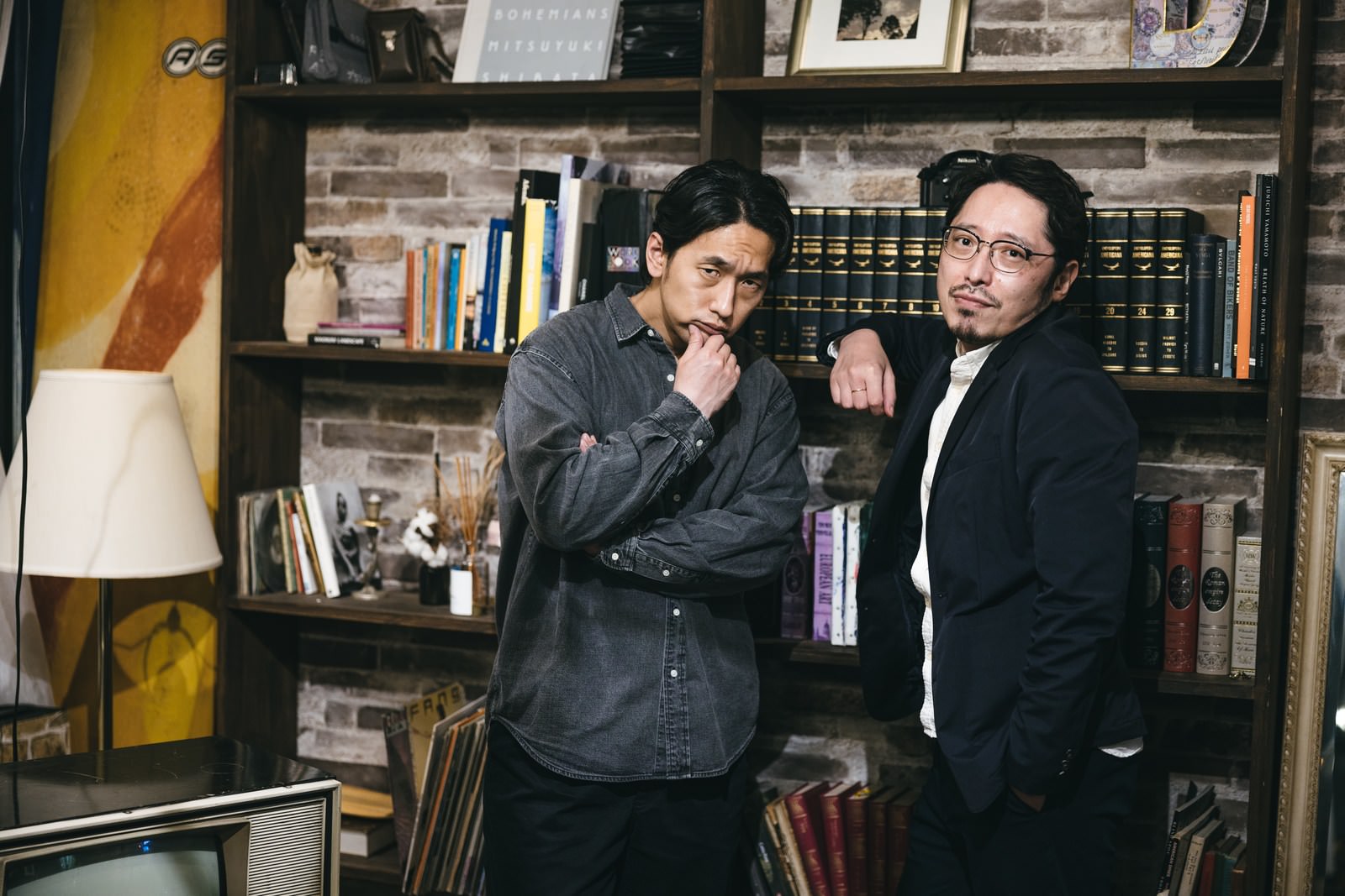 本棚の前で険しい表情を浮かべる黒い服装の男性二人組［モデル：大川竜弥 ゆうせい］