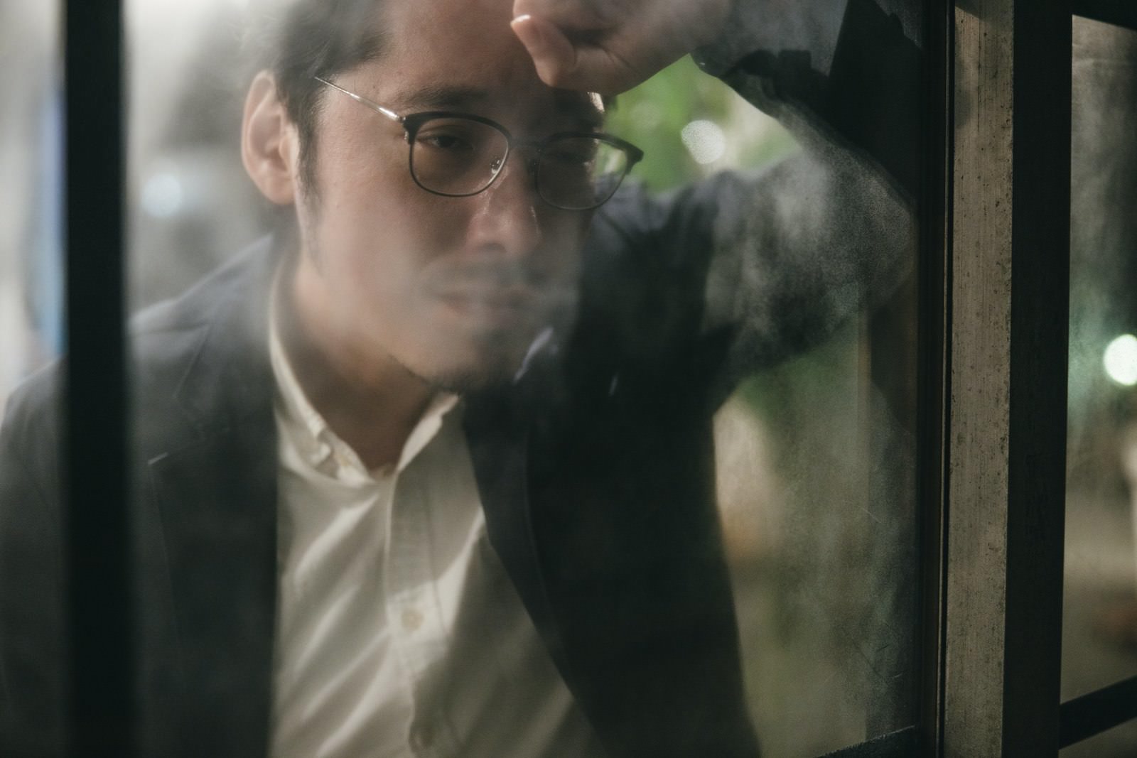 窓ガラス越しに額に手をあててうつむく、眼鏡をかけた男性［モデル：ゆうせい］
