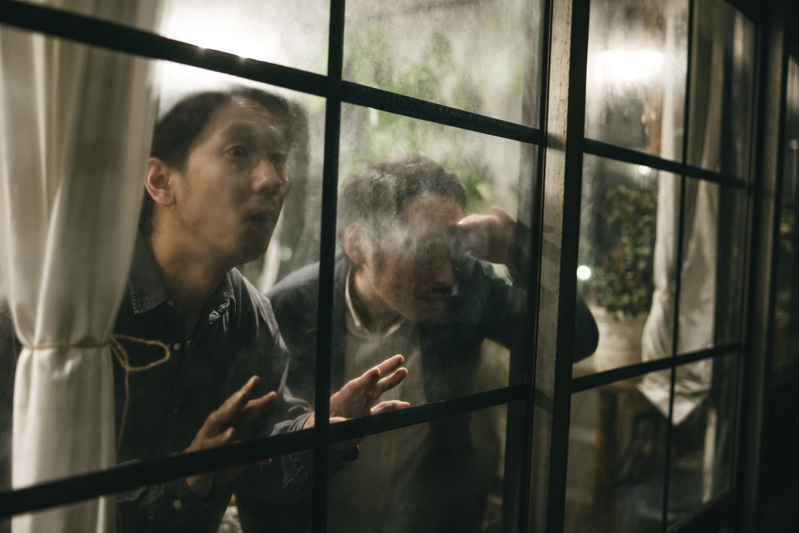 窓枠越しに外を見張る二人の男性が緊張感ある表情で張り込みをしている［モデル：大川竜弥］