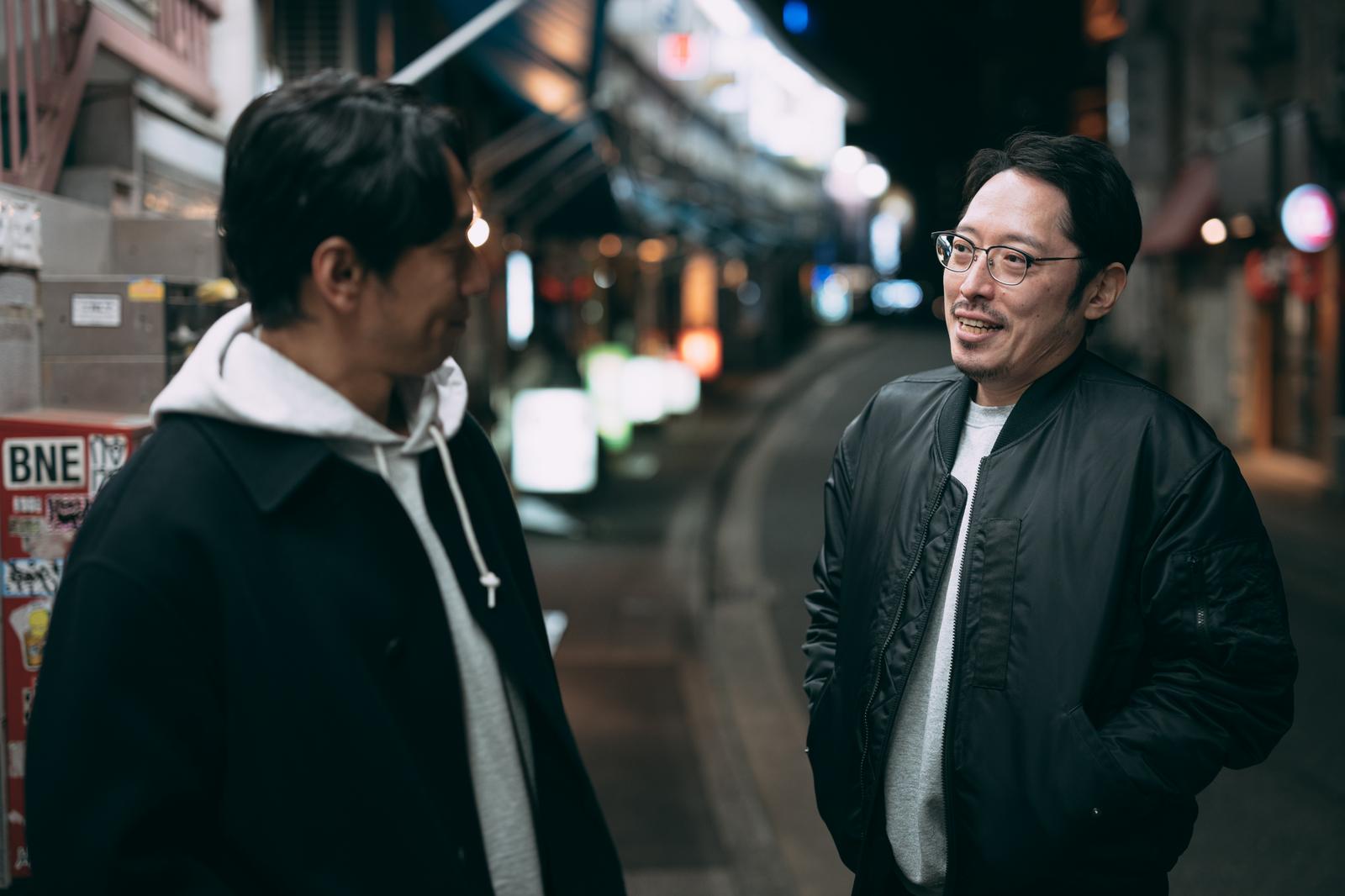 夜の街中で向き合って会話する眼鏡をかけた男性と別の男性［モデル：大川竜弥 ゆうせい］