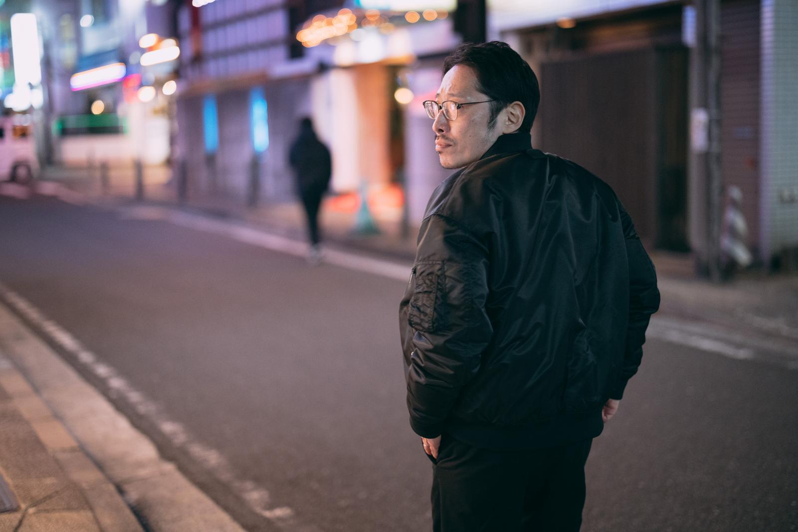眼鏡をかけた男性が夜間の繁華街で後ろ姿で立つ［モデル：ゆうせい］