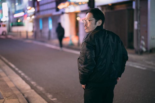 夜の街を歩く追跡者の後ろ姿、眼鏡の男性が黒いジャケット姿で尾行調査