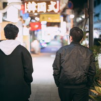 ネオンが輝く繁華街を夜間に歩く男性ふたりの後ろ姿の写真