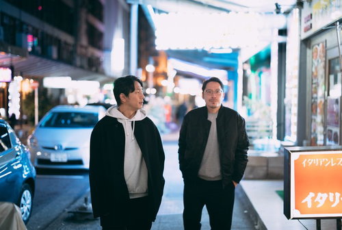 夜の繁華街で飲み屋を探す男性ふたり、ネオン照明の下町商店街を歩く