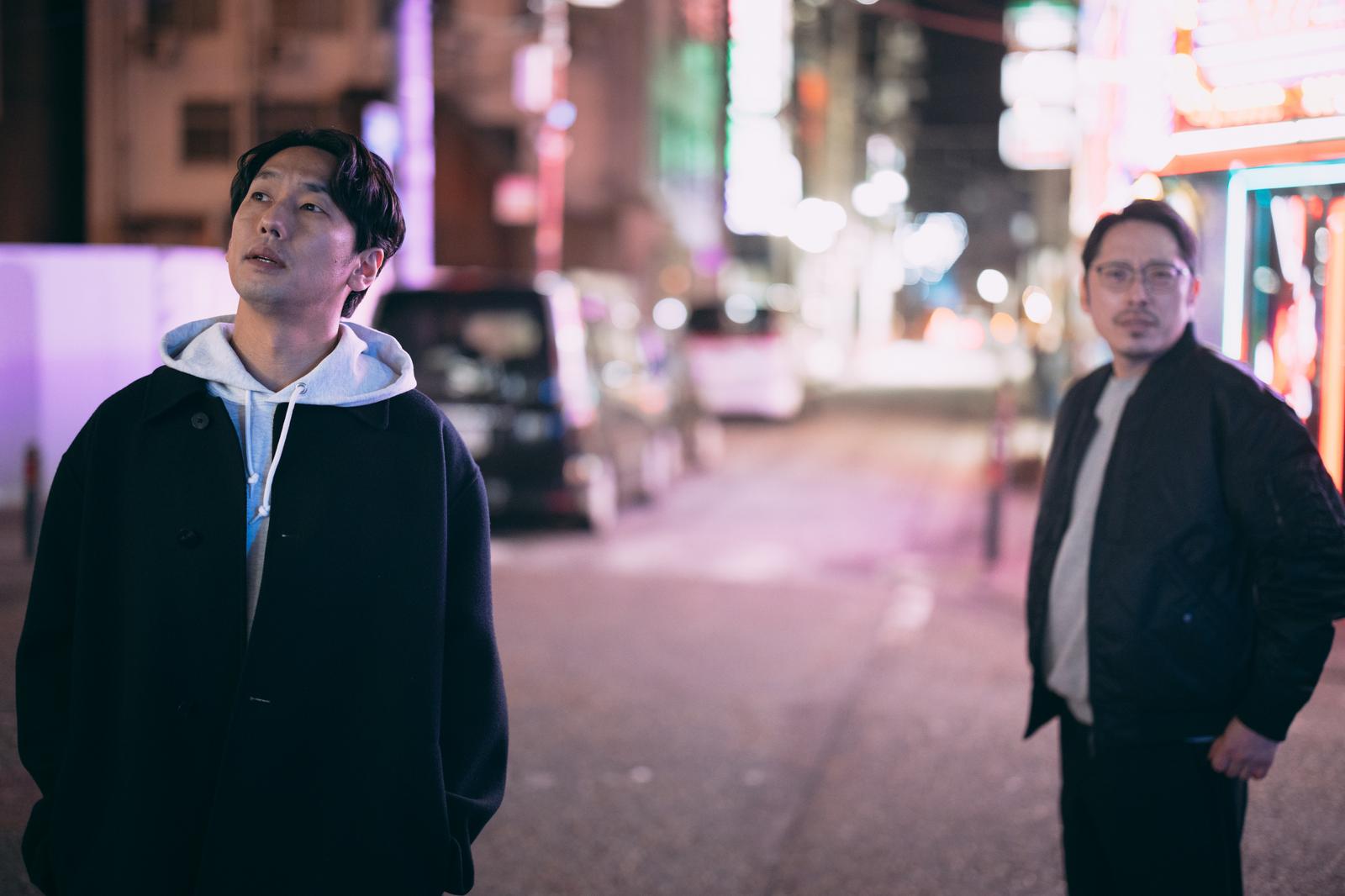 夜の街の繁華街で調査中の探偵二人の写真。ネオン看板が点灯する背景で、黒いフーディーの男性と眼鏡をかけた男性が立つ［モデル：大川竜弥 ゆうせい］