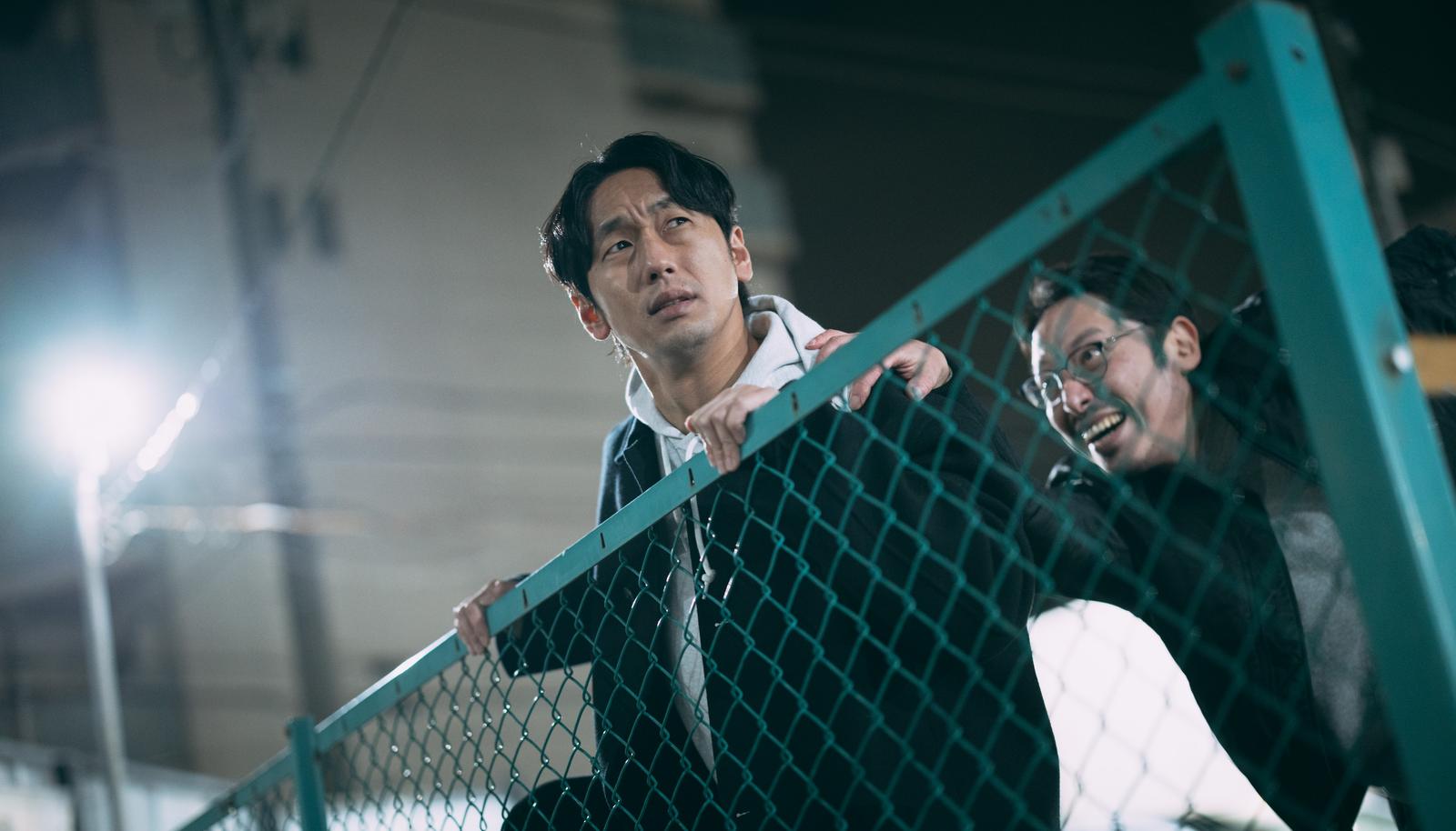 フェンス越しに逃げる白いシャツの男性と追いかける眼鏡姿の男性［モデル：大川竜弥 ゆうせい］