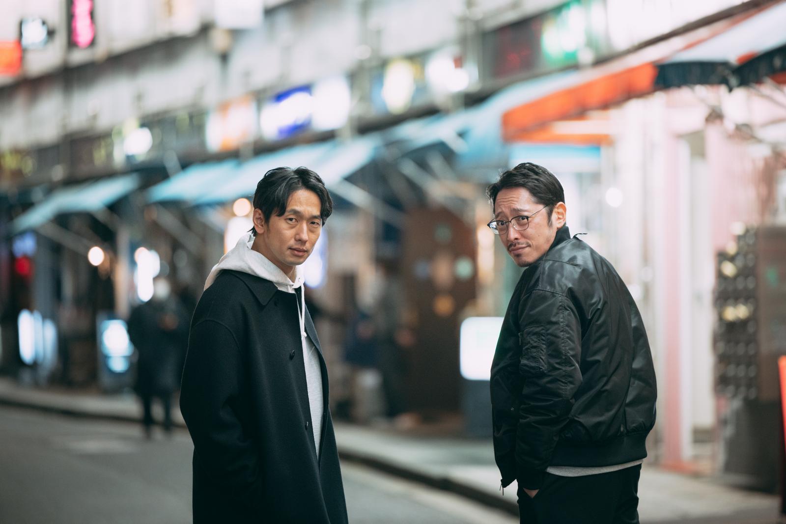 夜の駅前で立ち止まる黒いジャケット姿の二人の男性［モデル：大川竜弥 ゆうせい］
